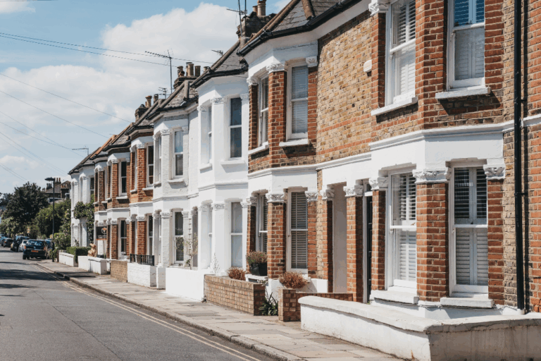 A row of traditional red-brick terraced houses with white-painted facades and bay windows on a quiet residential street in the UK, under a blue sky.