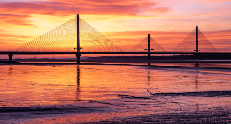 Cable-stayed Widnes Mersey Gateway Bridge over the River Mersey during a vibrant orange and pink sunset.