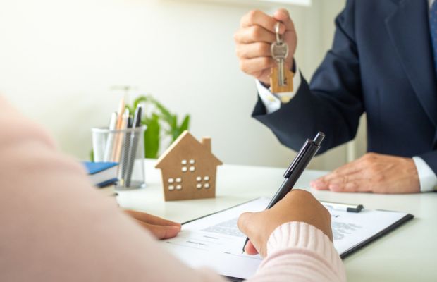 Person signing document; keys and small house model visible.