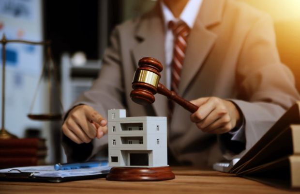 A person in a suit holds a gavel above a model house on a desk, with legal documents, books, and scales of justice visible in the background.