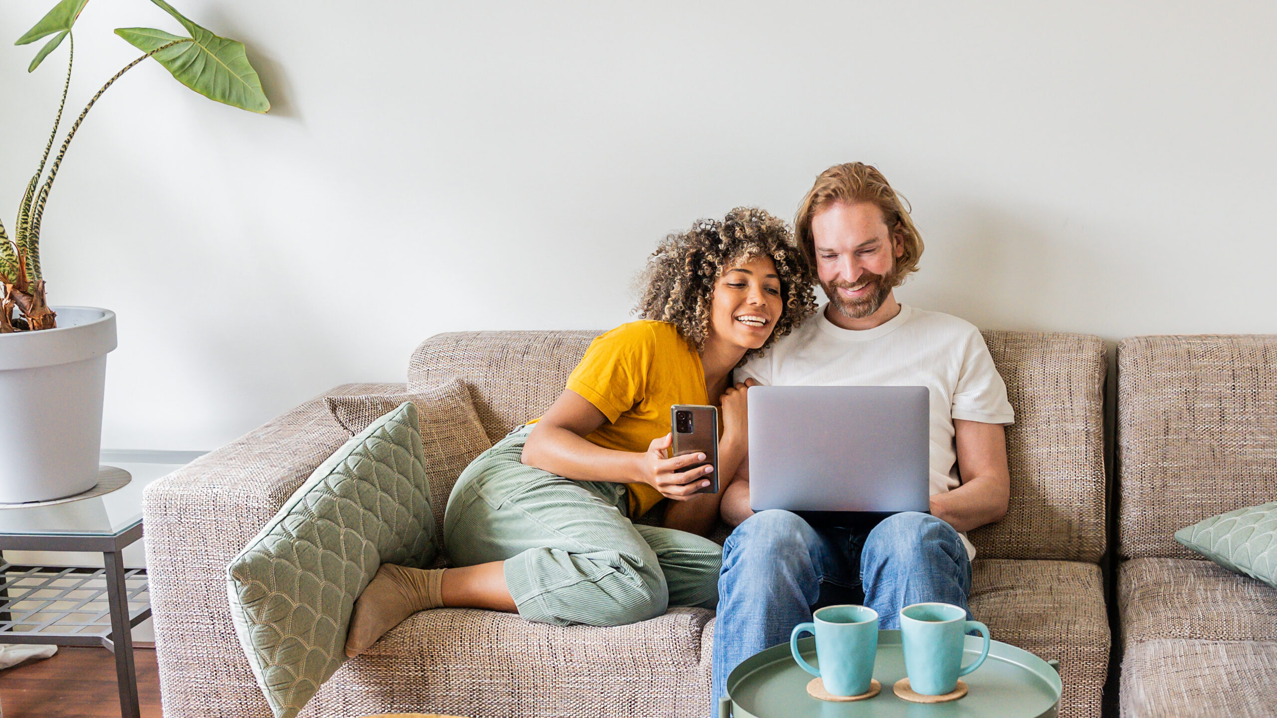 Couple sitting on a sofa using a laptop and phone while researching property, reflecting the process of buying a home in Ayr and understanding the Scottish market.