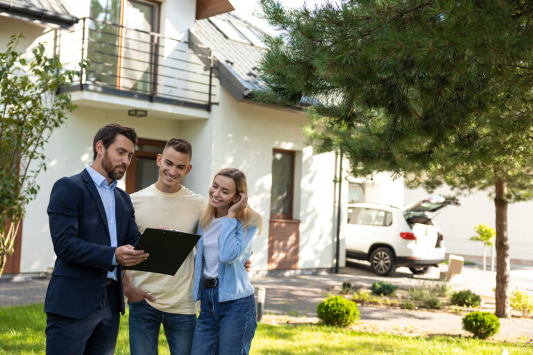 Estate agent reviewing a clipboard with a couple outside a modern house with a balcony and landscaped front garden.