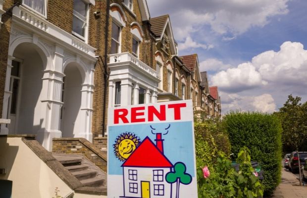 A For Rent sign stands in front of a row of residential houses, indicating available rental properties.