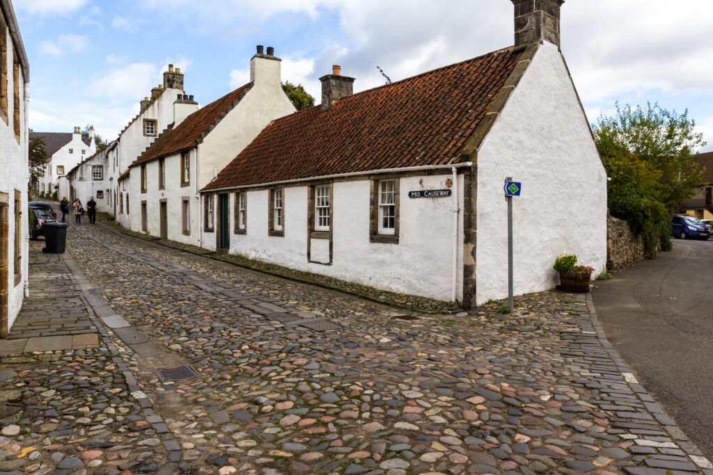 Whitewashed cottages with red clay tile roofs, stone chimneys, and narrow windows line a cobblestone street beneath a partly cloudy sky.