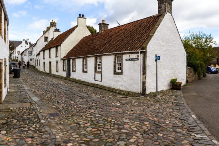 Whitewashed cottages with red clay tile roofs, stone chimneys, and narrow windows line a cobblestone street beneath a partly cloudy sky.