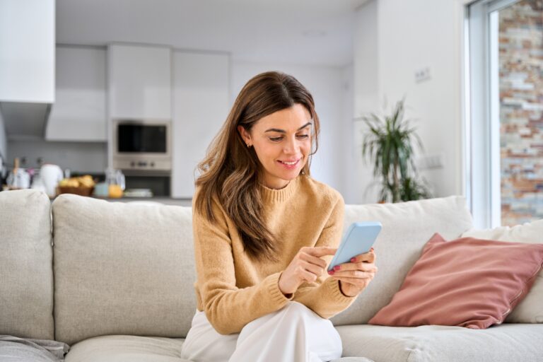 Smiling woman using her phone on a cream sofa in a bright open-plan home, representing living in Newton on Ayr KA8