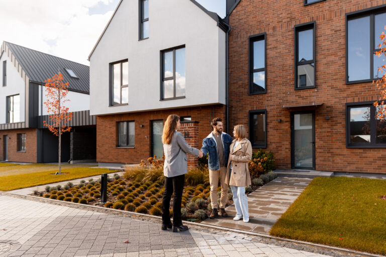 A real estate agent shakes hands with a smiling man and his partner outside a modern brick-and-white residential building with landscaped gardens